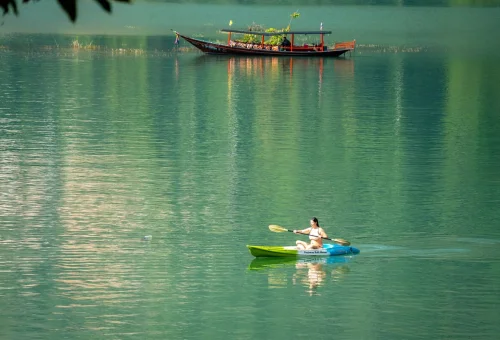 Visitor paddling a kayak across the emerald waters of Cheow Lan Lake with a traditional Thai longtail boat in the background