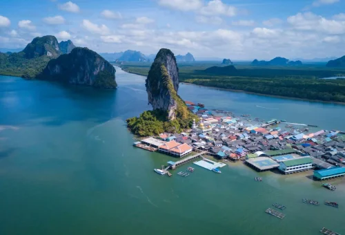 Drone view of Koh Panyee floating village — stilt houses, mosque, and football field surrounded by emerald waters and limestone cliffs, captured during the early departure James Bond Island Tour.