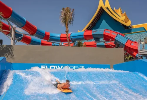 A guest riding an artificial wave on a surfboard at Andamanda Phuket Waterpark’s Wave Rider attraction, surrounded by cheering onlookers.