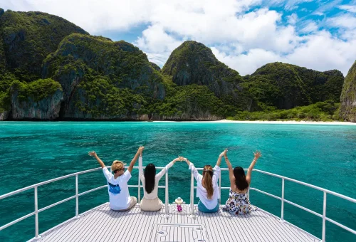 Guests on board a speed catamaran smiling and taking photos as they arrive at the iconic Maya Bay, with limestone cliffs and turquoise water in the background.