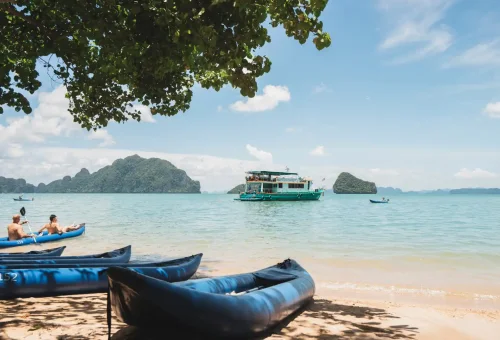 A tranquil beach scene with soft golden sand, gentle waves, and an Andaman sea kayak resting on the shore under tropical trees.