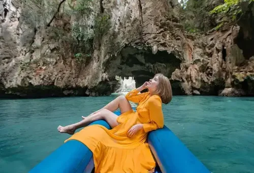 A woman posing in a canoe in front of a dramatic limestone cave, with calm emerald waters and a luxury catamaran anchored nearby at James Bond Island.