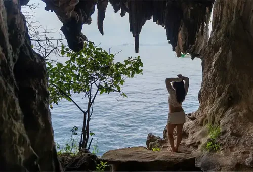 A woman standing at the edge of a sea cave, gazing out at the stunning cliffs and emerald waters of James Bond Island, with a luxury catamaran visible in the distance.