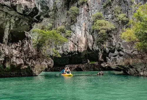 Canoes emerging from a limestone cave at Hong Island, with paddlers gliding into open waters near a luxury catamaran anchored nearby.