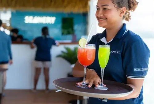 A smiling woman serving colorful cocktails on the deck of a party cruise boat in Phang Nga Bay, with festive vibes and scenic sea views in the background.