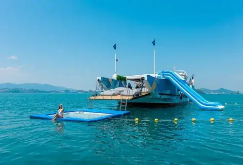 Guests enjoying a waterslide and floating water toys off the side of a party boat in Phang Nga Bay, surrounded by turquoise waters and limestone cliffs.