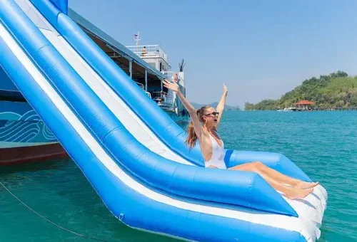 A woman laughing as she slides down a waterslide from a party boat into the clear waters of Phang Nga Bay, surrounded by tropical scenery.