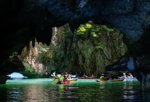 Guests canoeing through a sea cave at Hong Island during a stop on the Phang Nga Bay Party Cruise, surrounded by towering limestone cliffs and calm waters.