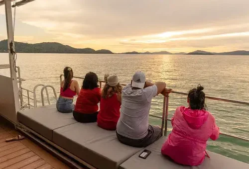 Customers relaxing and enjoying the golden sunset from the deck of a party boat in Phang Nga Bay, with vibrant skies and calm sea views.