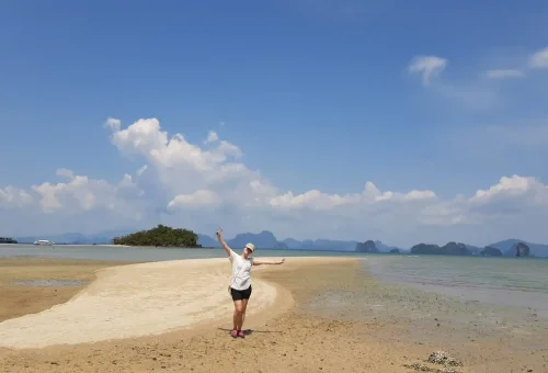 Cyclist standing with their bike on a quiet sandy beach at Koh Yao Noi, overlooking calm blue waters and distant islands under a sunny sky.