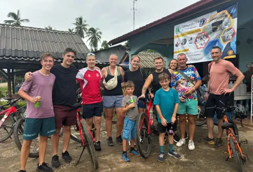 Cycling group posing for a photo in a peaceful village on Koh Yao Noi, surrounded by traditional wooden houses, palm trees, and smiling locals.