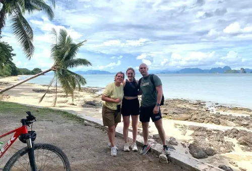 Cycling couple posing for a photo on a quiet beach at Koh Yao Noi, with bicycles nearby and a stunning view of the ocean and distant islands.