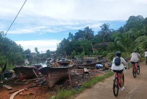 Cyclist riding through a traditional fishing village on Koh Yao Noi, passing wooden houses, long-tail boats, and coastal scenery.