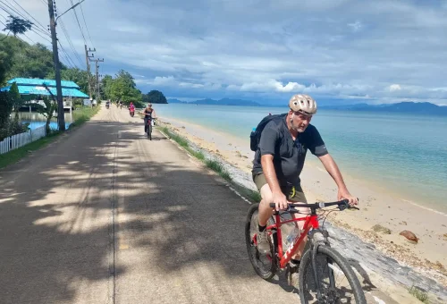 Cyclist riding along a scenic coastal road at Koh Yao Noi, with ocean views, palm trees, and distant limestone islands under a clear blue sky