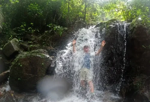 Traveler standing under a cascading jungle waterfall in Phuket, enjoying a natural shower surrounded by lush tropical rainforest.