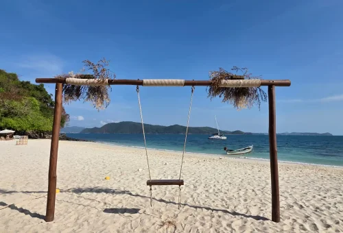 Guest enjoying a relaxing moment on a wooden swing set under palm trees at Banana Beach, Coral Island, with the ocean in the background