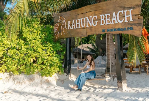 Scenic entrance sign "Welcome to Kahung Beach" with tropical surroundings, palm trees, and sandy pathway on Coral Island