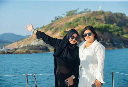 Two ladies posing at the bow of a sailing yacht with Promthep Cape in the background during a sunset cruise.