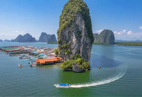 Aerial view of a speedboat approaching Koh Panyee floating village, surrounded by turquoise water and limestone cliffs in Phang Nga Bay
