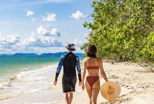 A couple walking hand in hand along the white sandy beach of Rang Yai Island, with turquoise water and palm trees in the background during the James Bond Island tour