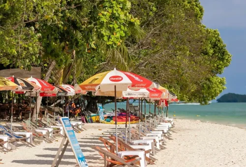 Beach chairs lined up on the white sandy shore of Koh Rang Yai with clear blue sea and tropical trees – PhuketTravelStore.com