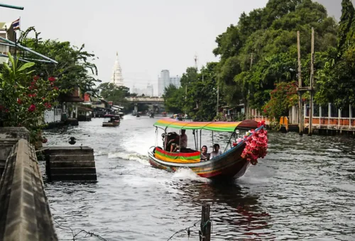 Longtail boat with tourists cruising along a wide Bangkok canal with city skyline and temple in the distance
