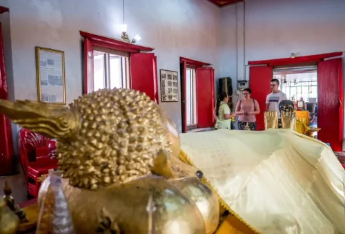 Interior of a Thonburi temple featuring a reclining golden Buddha statue with visitors in the background