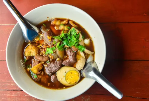 Bowl of traditional Thai stew with herbs, tofu, egg, and pork offal in a rich dark broth