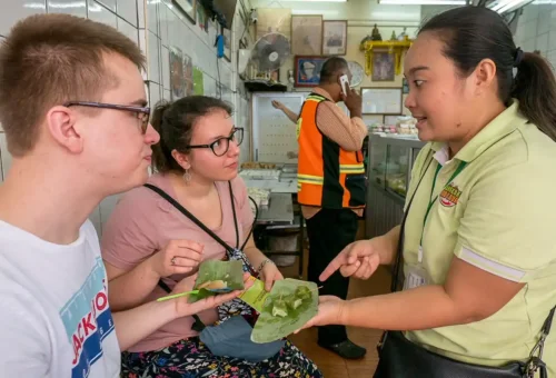 Local tour guide offering banana leaf-wrapped Thai snacks to curious tourists in Bangkok