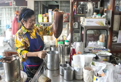 Female vendor in colorful shirt preparing traditional Thai coffee using a cloth strainer