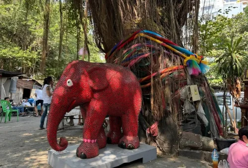 Red mosaic elephant statue under sacred banyan tree in Bangkok’s local neighborhood.