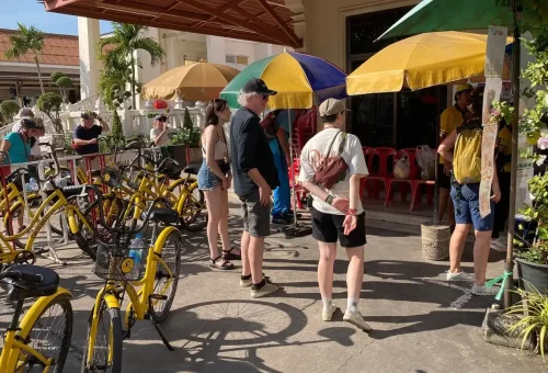 Travelers preparing for a Bangkok full-day bicycle tour under colorful umbrellas beside yellow rental bikes and a local temple.