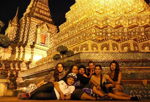 Happy cyclists resting at Wat Pho, marveling at the beauty of Bangkok’s ancient architecture lit up at night.