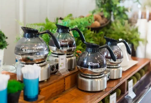 Tea and coffee dispensers arranged on a wooden buffet counter with greenery decor