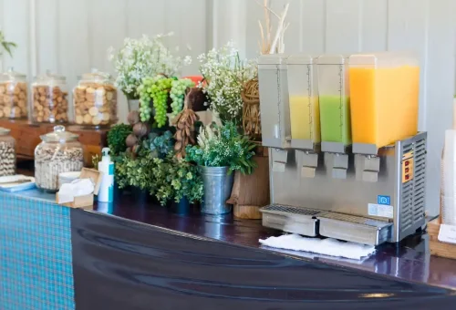 Beverage dispenser with orange, green, and yellow juices next to jars of snacks and decorative greenery