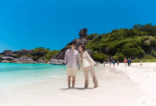 Smiling couple walking along the soft white sand beach at Similan Islands with Sail Rock in the background.
