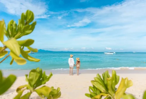 Couple holding hands on the beach looking out at the speedboat and sea.