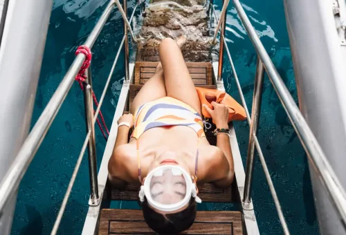 Woman relaxing on yacht stairs with snorkel gear above turquoise waters near Racha Island.
