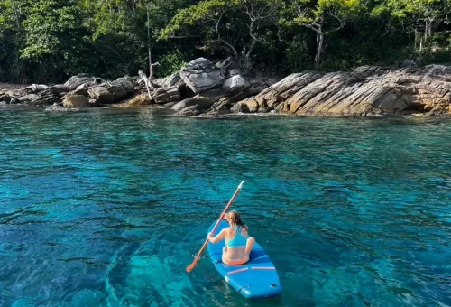 Solo paddleboarder near a rocky shoreline in turquoise waters by Racha Island.