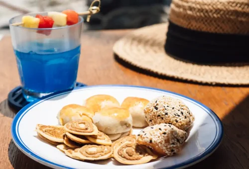 Plate of Asian pastries and rolls with a blue fruit drink, served on deck beside a sun hat.