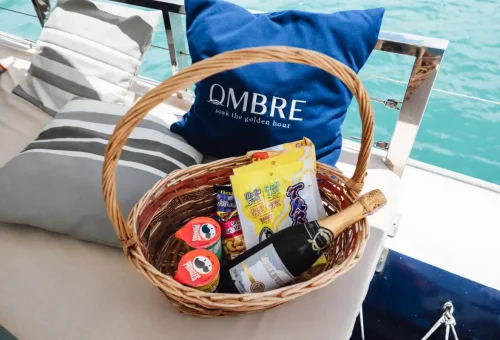 Picnic basket with wine, chips, nuts, and branded Ombre pillows on luxury yacht seating.