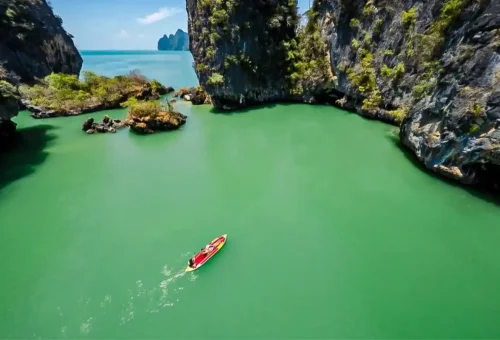 Red kayak paddling through emerald lagoon surrounded by limestone cliffs on Phang Nga Bay Sunset Serenity Cruise with Phuket Travel Store.