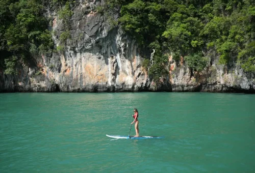 Woman standing on paddleboard in emerald waters near limestone cliffs on Phang Nga Bay Sunset Serenity Cruise with Phuket Travel Store.