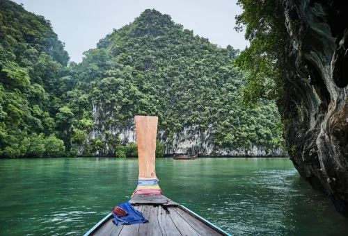 Longtail boat gliding through emerald waters surrounded by limestone cliffs and lush rainforest at Koh Hong, Krabi.