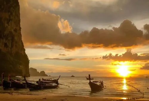 Sunset over Railay Beach with traditional longtail boats anchored in the sea.