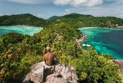 Traveler sitting on a rock viewpoint overlooking Koh Tao’s lush green hills and turquoise twin bays.
