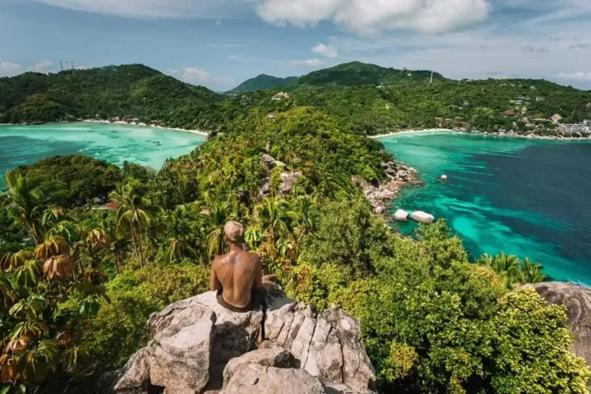 Traveler sitting on a rock viewpoint overlooking Koh Tao’s lush green hills and turquoise twin bays.