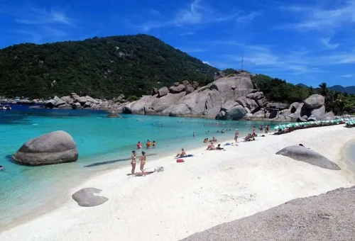 Tourists relaxing on the white sand beach of Koh Nangyuan with turquoise water and rocky cliffs in the background.