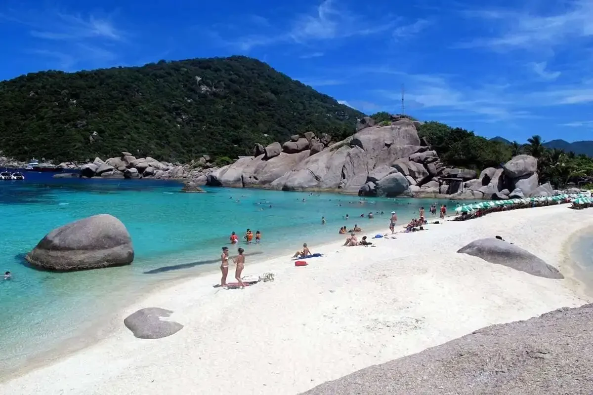Tourists relaxing on the white sand beach of Koh Nangyuan with turquoise water and rocky cliffs in the background.