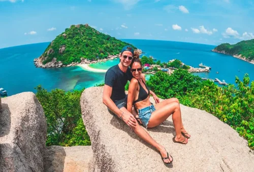 Happy couple sitting on rocks at Koh Nangyuan viewpoint overlooking turquoise sea and white sandbar.